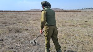 Using the metal detector on the field with a flak jacket, helmet, and safety glasses